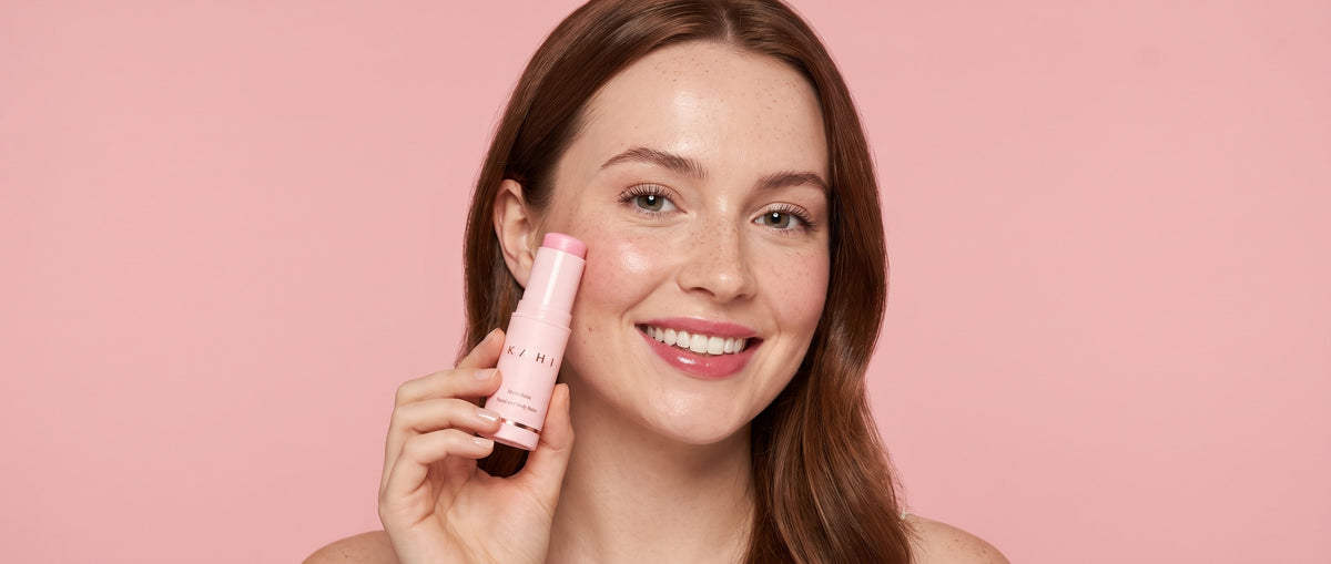 A young woman smiling as she holds the KAHI Multi Balm stick to her cheek